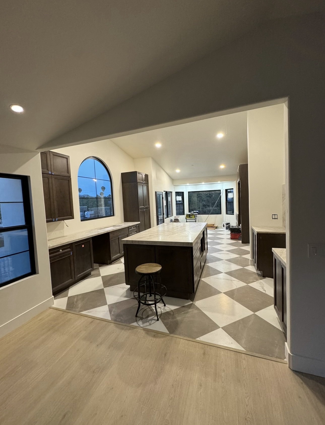 Wide view of a finished kitchen remodel in the Bonita / Chula Vista area with dark wood cabinetry, marble island and backsplash, pro range with brass pot filler, checkered tile floor, and arched black-framed window