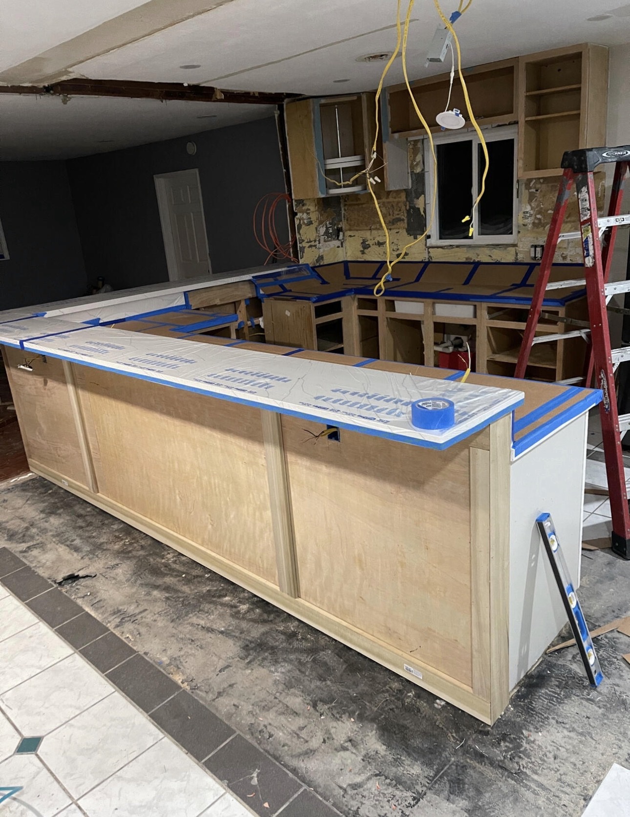 Open-concept kitchen remodel during cabinet installation with a new island base, protected white counters and electrical rough-in overhead