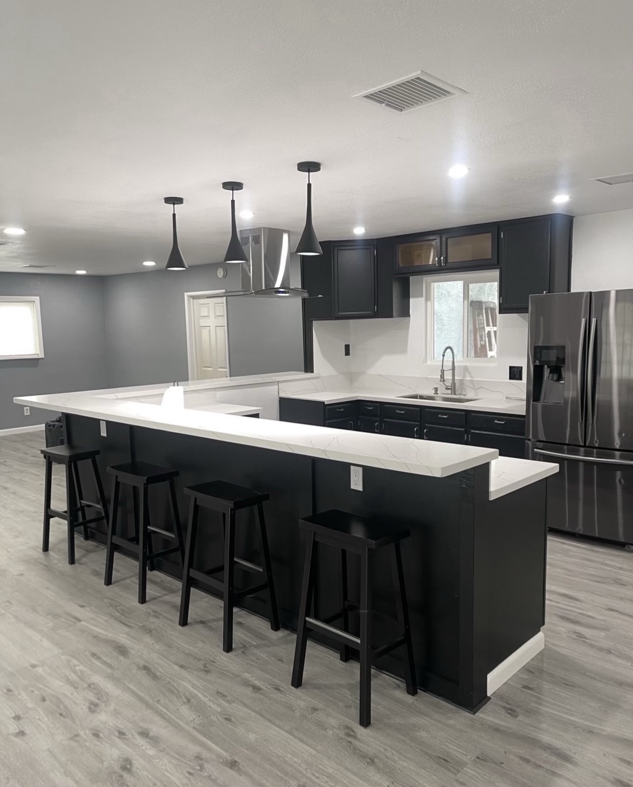 Finished open-concept family kitchen with black cabinets, white marble-look island, bar stools, stainless steel refrigerator and gray wood-look flooring